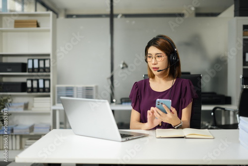 Serious call center operator in wireless headset talking to customer, woman in headphones with microphone consulting customer on phone in customer support service, looking at computer screen.