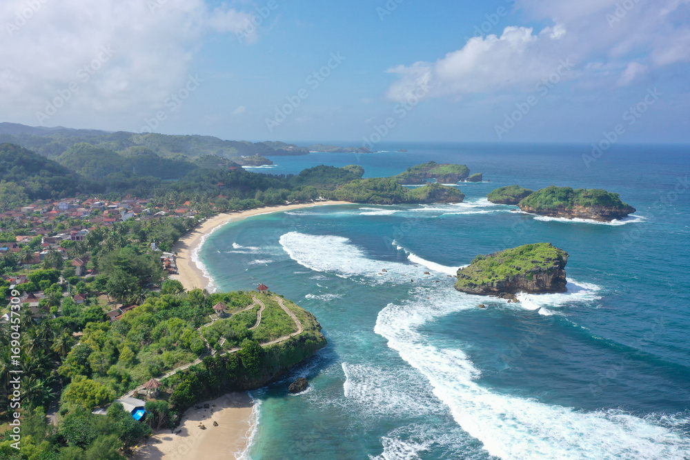 Fototapeta premium Aerial drone view of a scenic tropical coastline featuring a sandy beach, a village nestled among green hills, and several rocky islands in the vibrant blue ocean. A perfect travel shot.watu karung