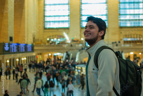 Smiling young Hispanic backpacker inside Grand Central Terminal in New York City surrounded by commuters and tourists enjoying lifestyle, architecture and urban tourism in iconic station