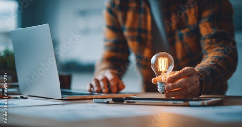 A person in a plaid shirt holds a glowing lightbulb at a desk with a laptop and tablet. Focus is on the bulb and hand