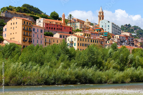 A view of a beautiful Italian old town Vintimille rising up into the hillside towards mountains in the distance. There are painted houses and church spire and wooded hills.
