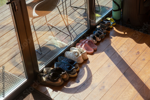 A row of shoes and sandals neatly arranged on a wooden floor by a glass door, illuminated by warm sunlight streaming inside.