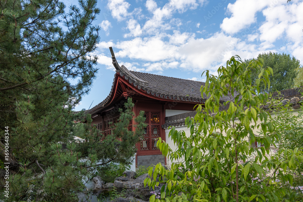 Fototapeta premium A red Chinese-style pavilion stands among trees and shrubs. Its curved roof stands out against the blue sky.