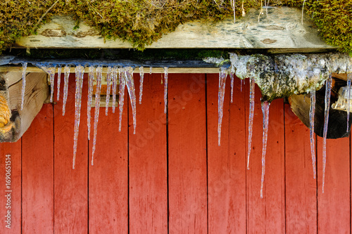 Icicles form under a weathered wooden eave, contrasting against a vibrant red wall, showcasing winter's chill and beauty.