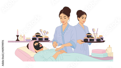 Two women in spa robes applying a black facial mask to a client lying on a massage table with beauty products around them