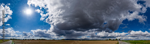 Panoramafoto einer Wolkenlücke mit der Sonne und dunkler Regenwolke im Gegenlicht über landwirtschaftlich genutzter Fläche mit zwei leeren Landstraßen