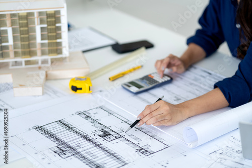 Asian adult architect professional carefully analyzes detailed building blueprints using calculator at office table