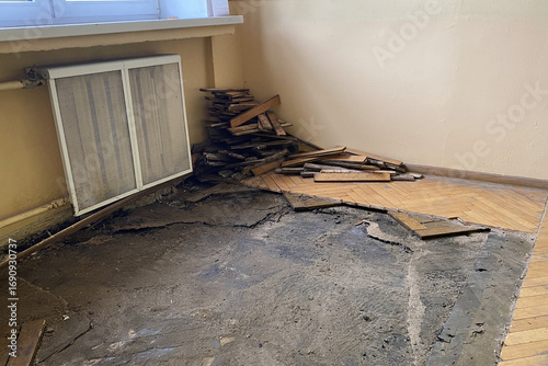 Room under renovation with partially removed parquet flooring, exposing concrete subfloor and stacked wooden planks in the corner.