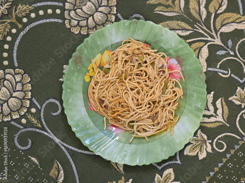 A plate of cooked noodles sits on a floral-patterned, green tablecloth. The green plate features decorative flowers and is filled with noodles mixed with sliced vegetable.