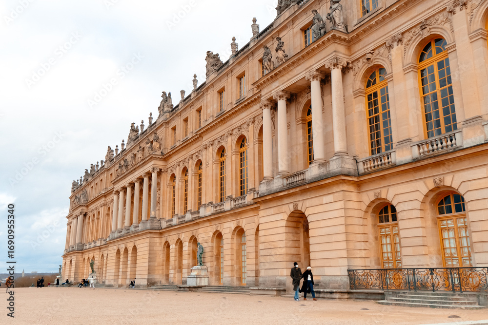Obraz premium Visitors admire the stunning facade of Palatul Versailles, marveling at its intricate architecture and historical significance under a cloudy sky.