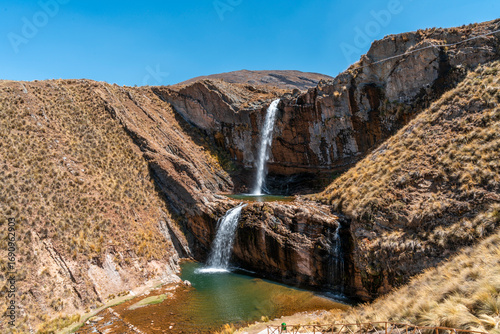Tourist view of a two-level waterfall in Marcapomacocha, Peru, cascading into turquoise pools surrounded by rocky cliffs and Andean highlands.