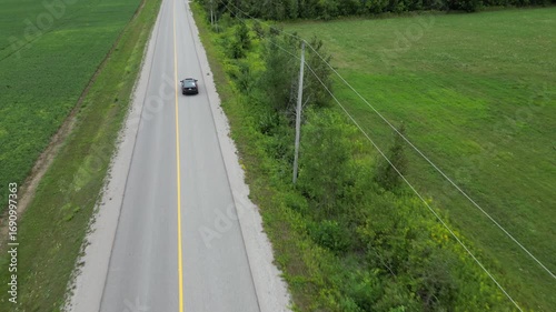 Aerial Drone footage of car driving along a rural road with green fields and trees