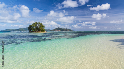 Small island with a tree , the Indian Ocean, blue sky with clouds and an island on the horizon. La Digue, the Seychelles