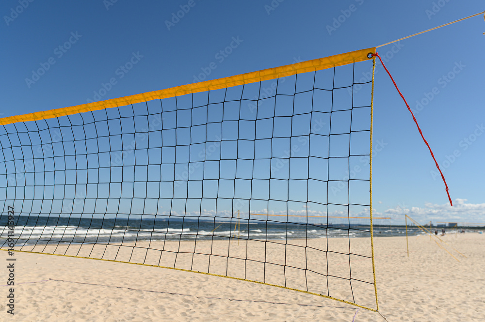 Fototapeta premium A vibrant beach volleyball net stands tall on warm sand, facing gentle ocean waves under a clear blue sky