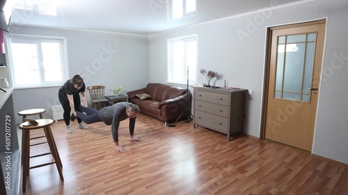 Holding partner legs for wheelbarrow walk, middle aged white woman in sportswear and mature man in homewear exercising together on wooden floor.