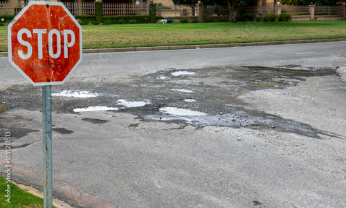Old looking stop sign on the left and water filled potholes in a damaged asphalt road surface in the background.  