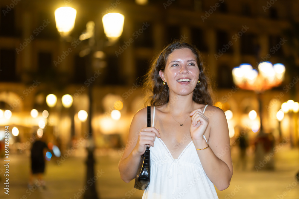Fototapeta premium Long-haired cheerful female brunette walks along city street, spends time in motion, walks and enjoys sunny warm day, walks from university to home. Carefree, night cafe, promenade.