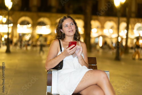 Canvas Print Woman is sitting on a chair after a long walk through Barcelona at night, she is resting and writing a message on her mobile