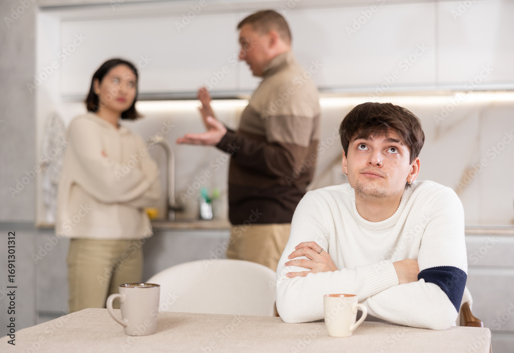 Fototapeta premium Young guy sad while sitting at table during family quarrel between adult man and young woman