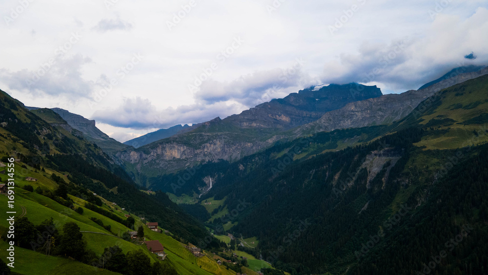Obraz premium Swiss Alps with houses, winding road, clouds, and overcast sky, captured by drone. Schweizer Alpen mit Häusern, kurviger Straße, Wolken und bedecktem Himmel, aufgenommen mit Drohne.