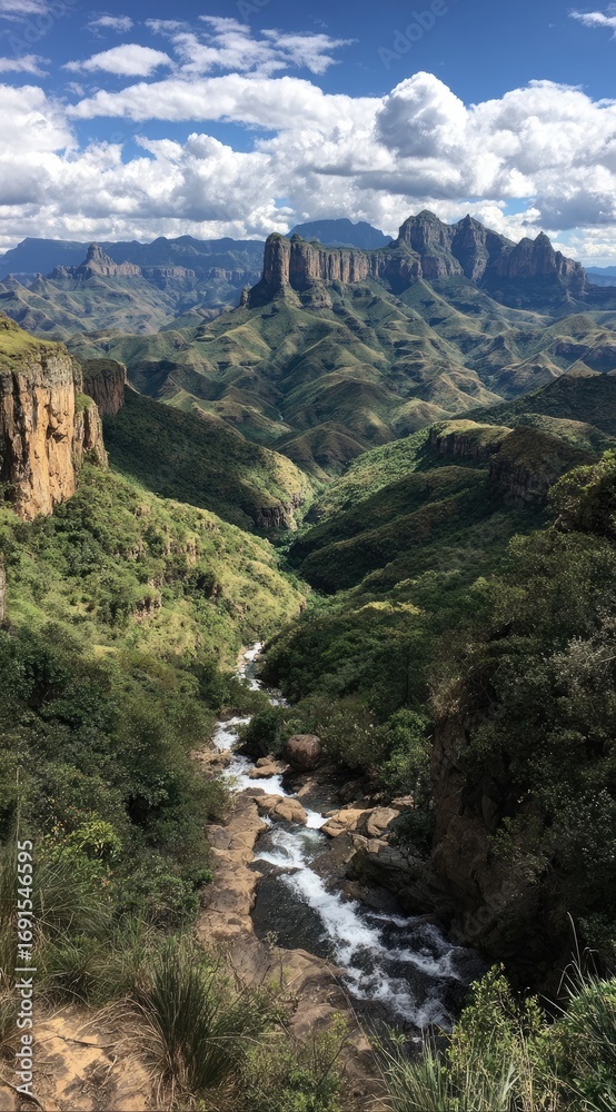 Fototapeta premium A meandering river carves a path through a lush, green valley, framed by towering cliffs and rugged mountains under a bright, cloudy sky