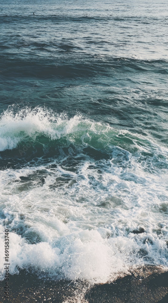 Fototapeta premium Seascape view features frothy waves crashing onto a dark shoreline. White foam contrasts the deep blue and turquoise of the ocean water under soft daylight