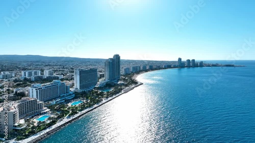 Aerial view of a modern coastal city with high-rise buildings lining a long beach and the clear blue Mediterranean Sea under a bright sky.