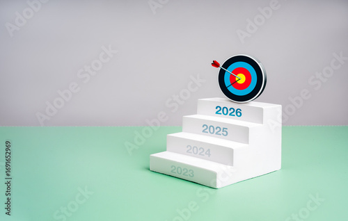 White staircase with years 2023 to 2026 leading to 3d colorful target hit by an arrow, symbolizing business growth, long-term planning, goal setting, and success vision for the new year 2026.