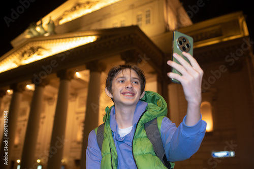 A teenage boy holds a smartphone in his hands, takes a selfie while standing in front of the Bolshoi Theater in the evening in Moscow.