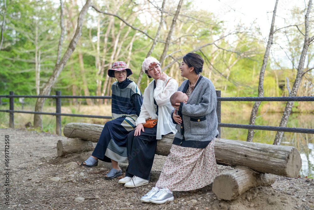 Fototapeta premium Three women sitting on a log in a park. One of them is holding a baby. Scene is peaceful and relaxing