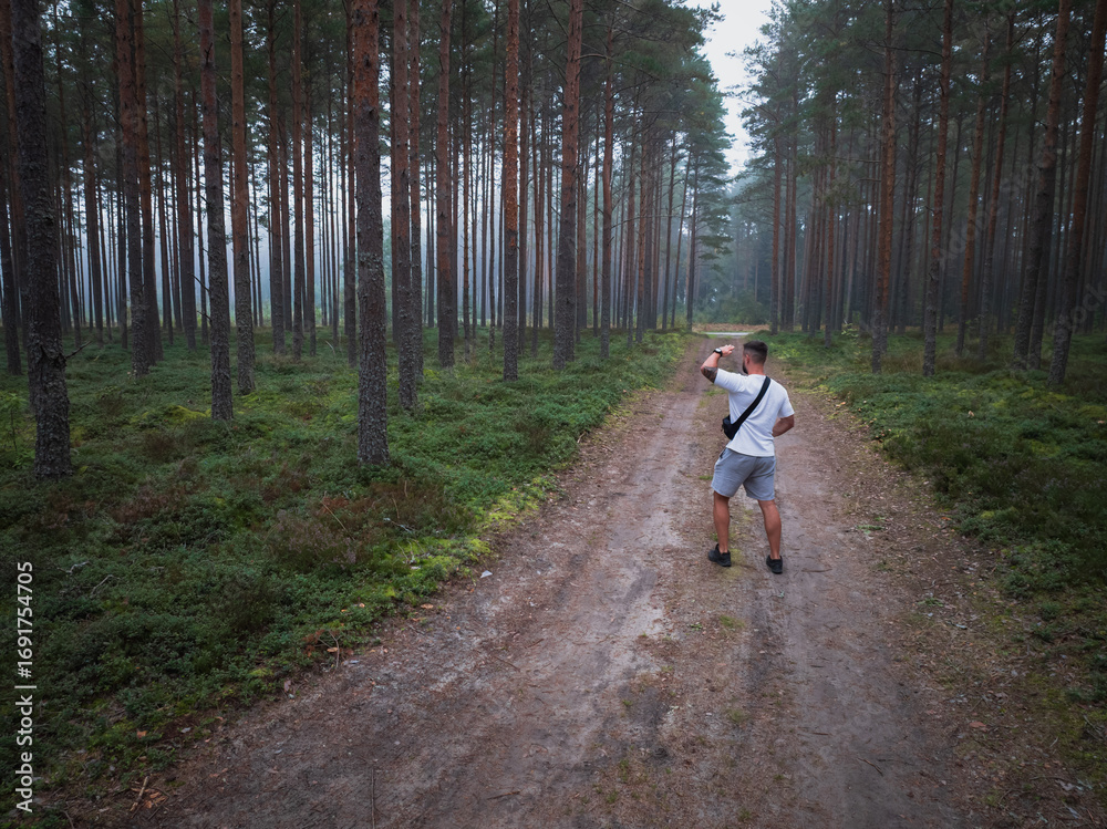 Fototapeta premium Aerial drone view of a man hiking on a dirt road through a misty pine forest, exploring the wilderness.