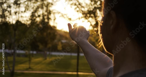Womans hand reaches out to sun in park, at golden sunset. Fingers spread as sunlight shines through. Scene shows freedom, hope and inspiration. Warm light, atmosphere of motivation and inner strength.
