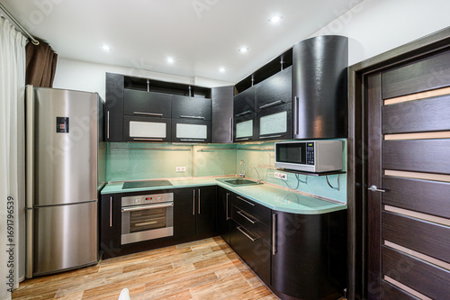 modern kitchen features dark wood cabinetry, a stainless steel refrigerator and oven, and light green countertops. Recessed lighting illuminates the space