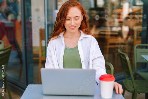 Young woman working on laptop at outdoor cafe enjoying coffee while smiling in bright daylight in a lively atmosphere