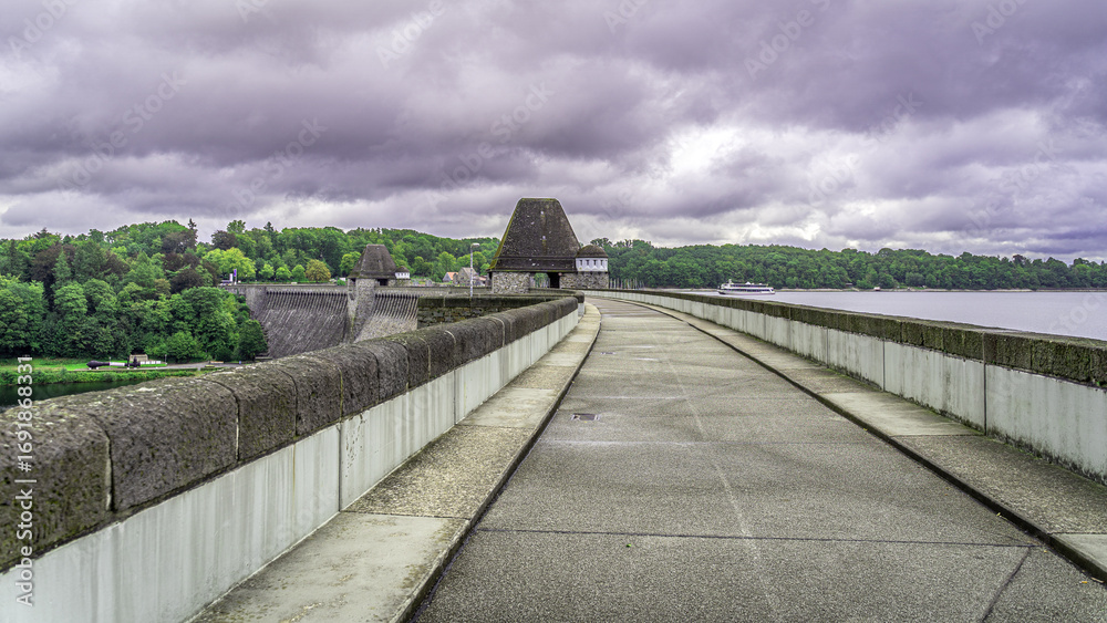 Fototapeta premium Der Weg über die Talsperre führt direkt in die Weite, unter einem dramatisch bewölkten Himmel. Die ruhige Oberfläche des Sees und das historische Bauwerk erzählen von der Kraft der Natur und der mensc