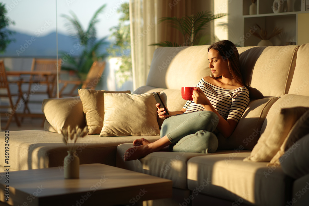 Obraz premium Woman drinking coffee using mobile phone sitting on a sofa