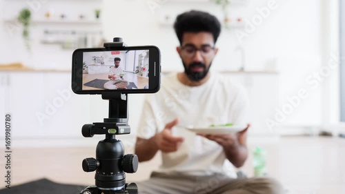 Young adult male with beard records video blog in home kitchen holding salad bowl, discussing healthy eating habits and sharing tips. Scene shows content creation, cooking, modern lifestyle concepts.