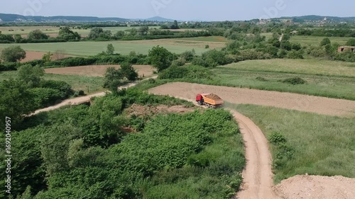 A bright orange dump truck drives along a rural dirt road surrounded by lush greenery and muddy construction terrain. The aerial view captures a contrast between nature and land development.