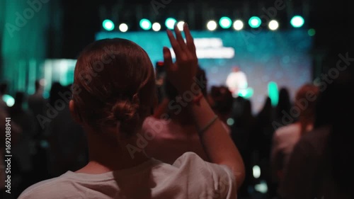 Female worshipper lifting hand during live music event, surrounded by crowd, large screen projecting colorful background visuals