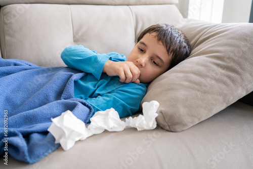 Sick child feeling unwell at home, coughing into fist while lying on couch under white blanket with tissues nearby, ill male kid resting during the day in quiet living room, closeup