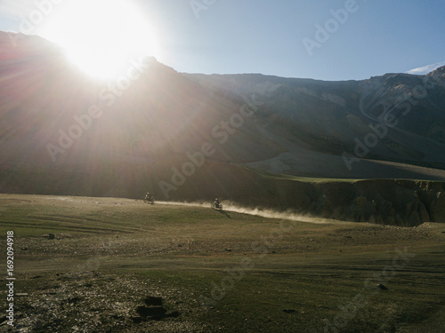 Aerial view of the sun blazing over the stark mountains, casting long shadows across the rugged terrain where motorbikes kick up dust, Kaza, Himachal Pradesh, India.