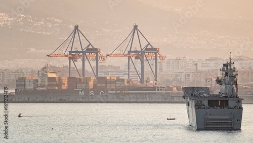 Military ship and container vessel in busy seaport at sunset, with giant cranes and city skyline in the background
