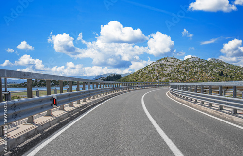 Scenic coastal highway with a curve and guardrails overlooking mountains, blue sky, and water on a bright summer day.