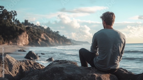 A man sits on a rock on a beach, looking out at the ocean. The sun is setting in the background, and there are clouds in the sky.