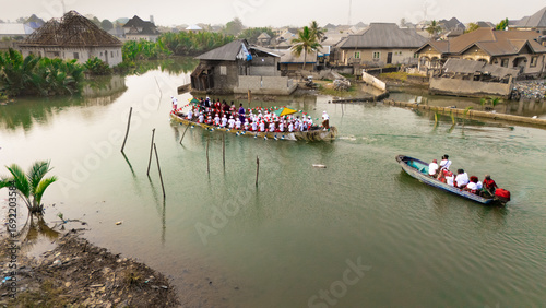 Aerial view of War Canoe of the Niger Delta carrying warriors navigate through waterways surrounded by houses, Ogu Town, Rivers State, Nigeria.
