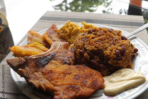A top view of a plate of delicious Gallo pinto and pork chop with plantain, scrambled egg, and custard on a wooden table, a typical dish of Costa Rica.