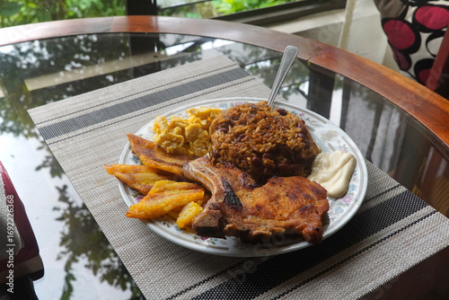 A top view of a plate of delicious Gallo pinto and pork chop with plantain, scrambled egg, and custard on a wooden table, a typical dish of Costa Rica.