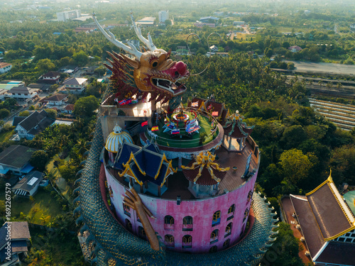 Aerial view of the vivid pink Wat Samphran temple entwined by a colossal dragon, its scales shimmering against the lush green landscape, Wat Sam Phran, Bangkok, Thailand.