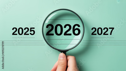 Conceptual image with years 2025, 2026, 2027 in a row on pastel green background. A hand holds a magnifying glass over 2026, enlarging it to symbolize focus, planning, and reflection