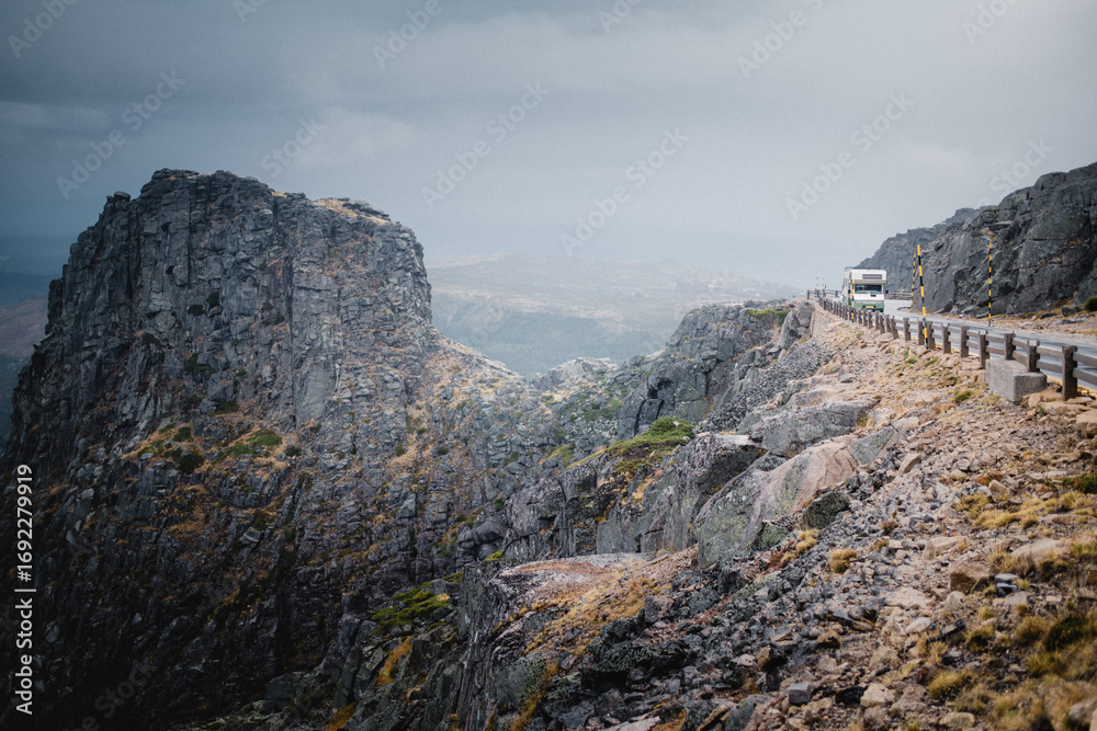 Fototapeta premium A winding mountain road in Serra da Estrela under gloomy weather, with dark skies pressing over the rugged landscape. The scene feels dramatic, remote, and atmospheric.